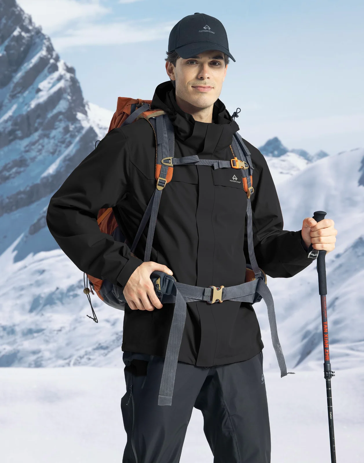 A man on a snow slope in a black insulated jacket, a pack on his shoulders and a pole in each hand, mountains behind him